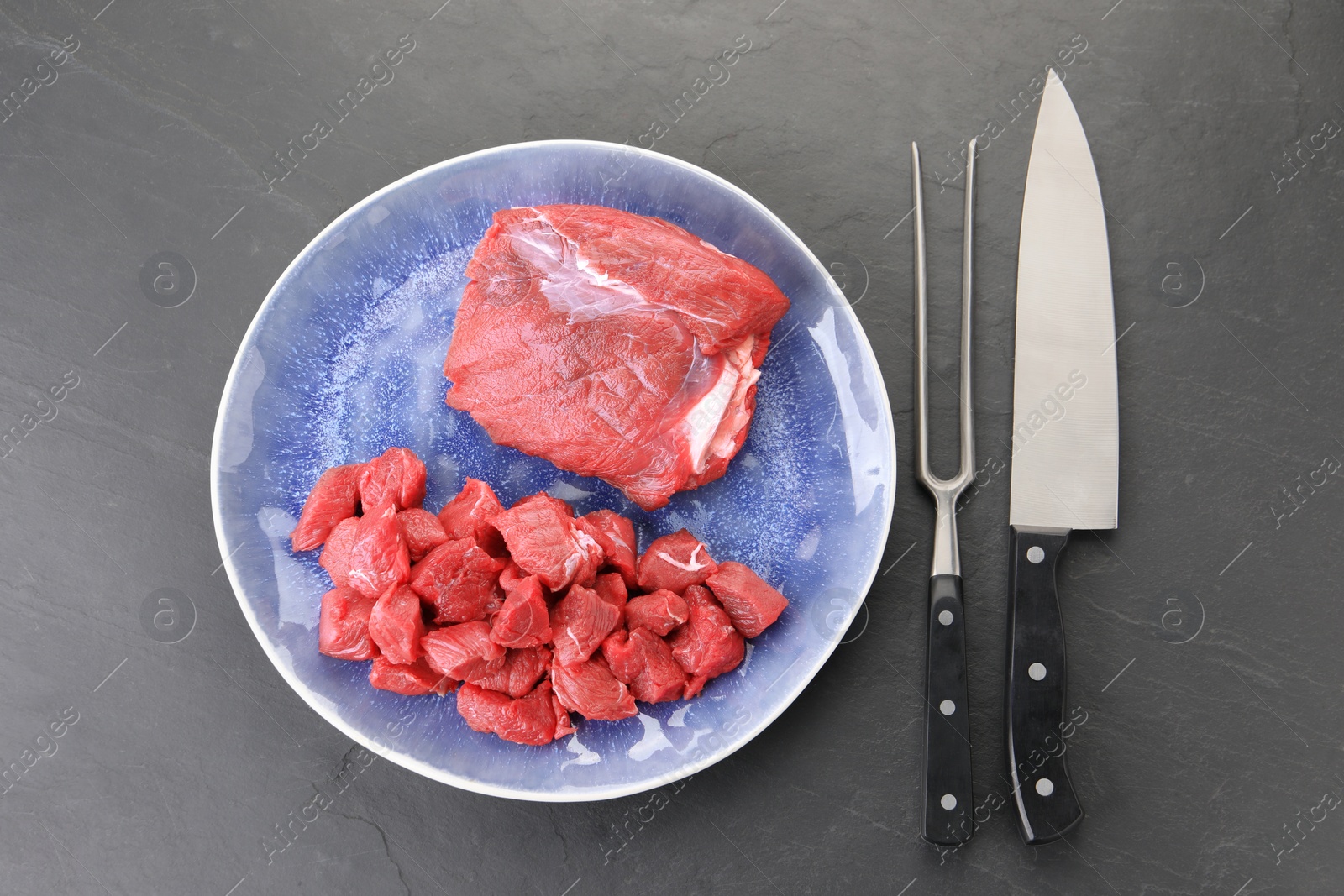 Photo of Plate with pieces of raw beef meat, knife and fork on grey textured table, flat lay