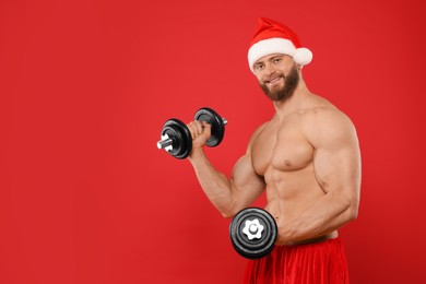 Muscular young man in Santa hat with dumbbells on red background, space for text
