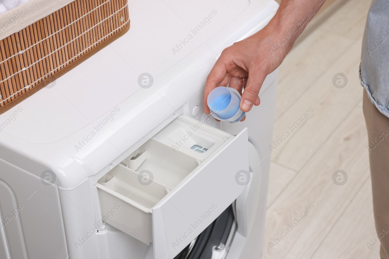 Photo of Man pouring fabric softener from cap into washing machine indoors, closeup