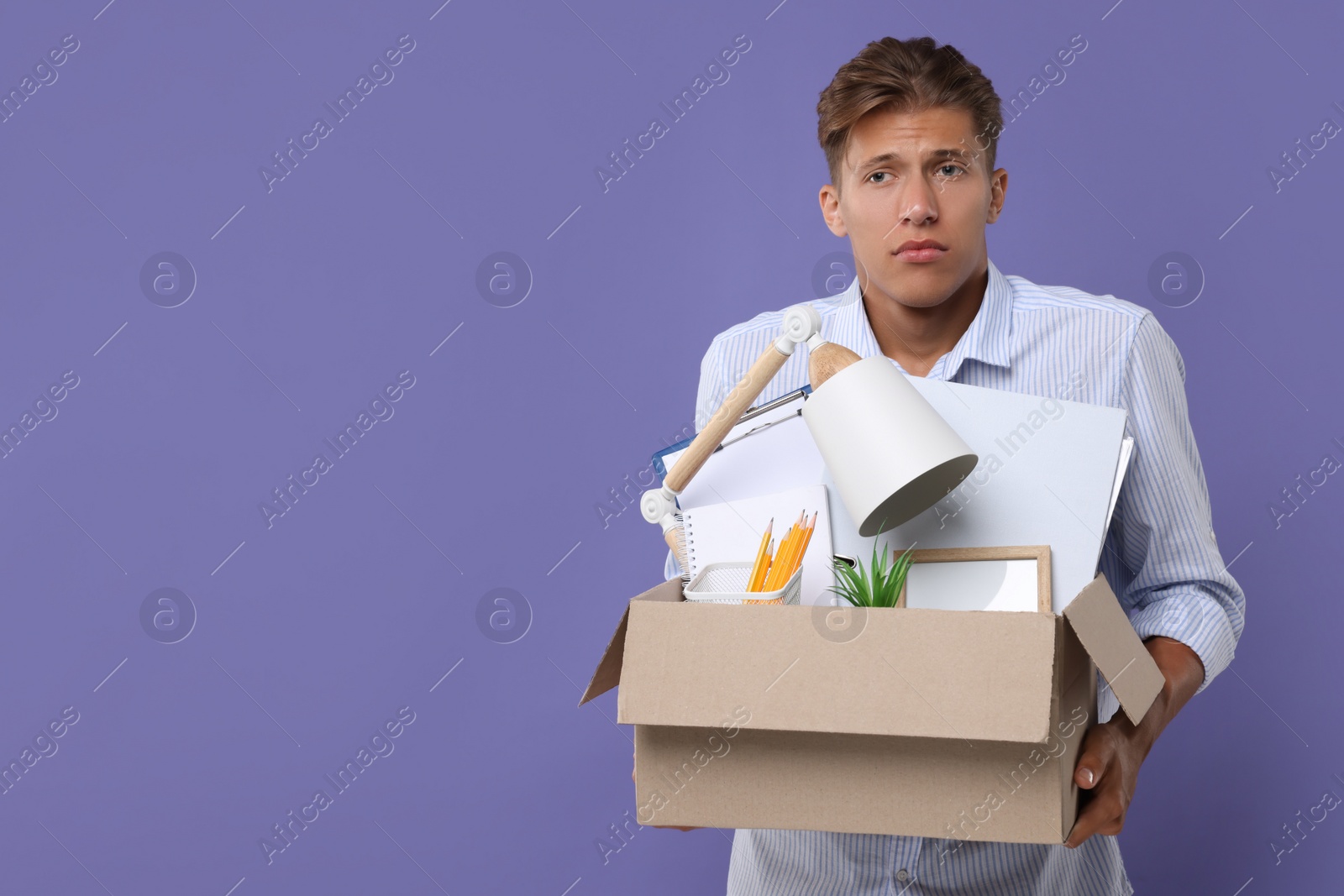 Photo of Unemployed young man with box of personal office belongings on purple background. Space for text