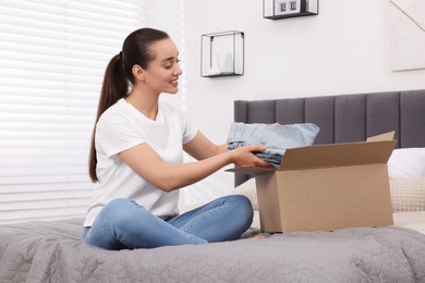 Photo of Happy young woman taking jeans out of box on bed at home. Online shopping