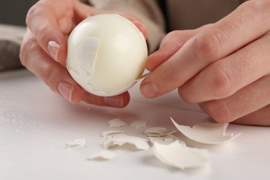 Woman peeling boiled egg at white wooden table, closeup