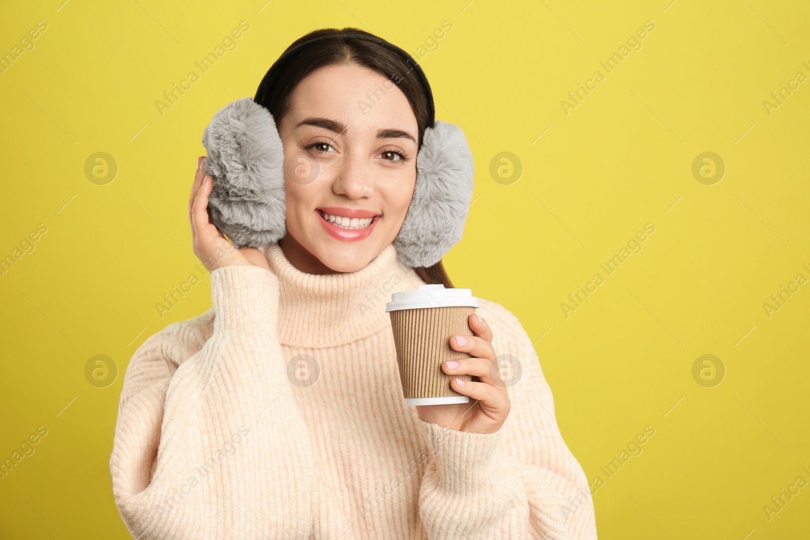 Photo of Beautiful young woman in earmuffs with cup of drink on yellow background. Space for text