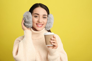 Photo of Beautiful young woman in earmuffs with cup of drink on yellow background. Space for text