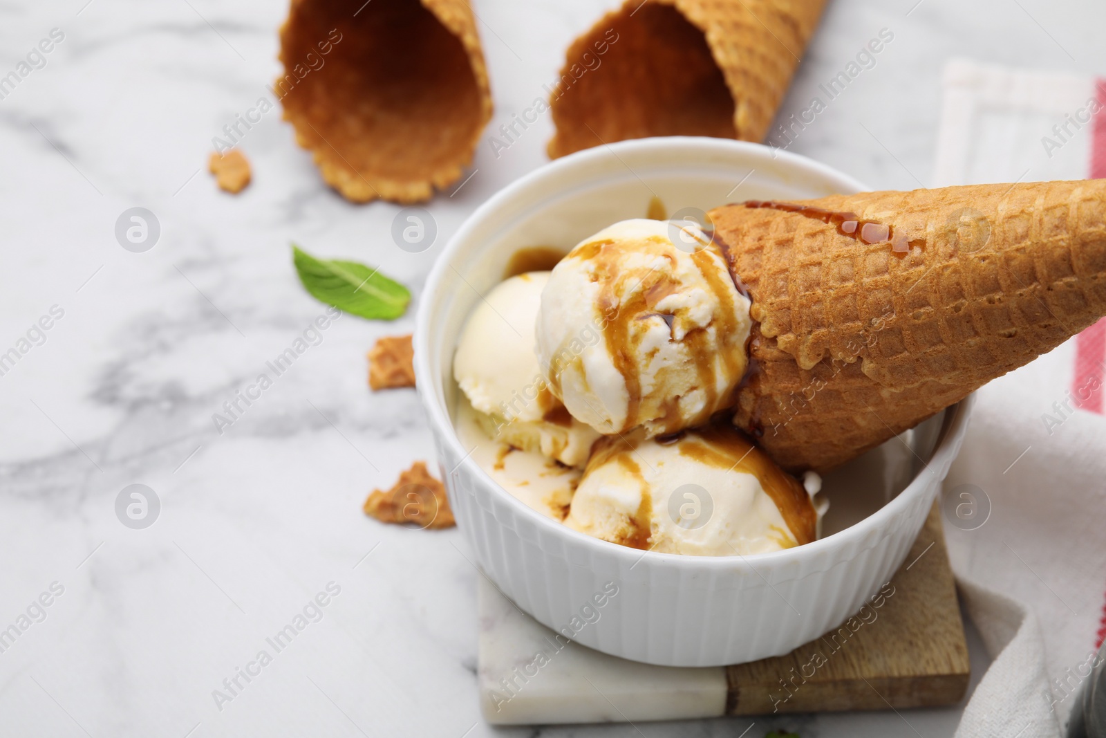 Photo of Scoops of ice cream with caramel sauce and cone on white marble table, closeup. Space for text