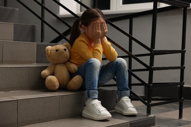 Photo of Child abuse. Upset little girl with teddy bear sitting on stairs indoors