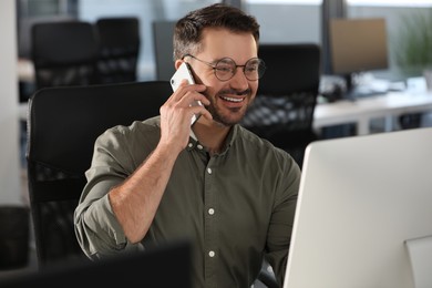 Happy man using modern computer while talking on smartphone in office