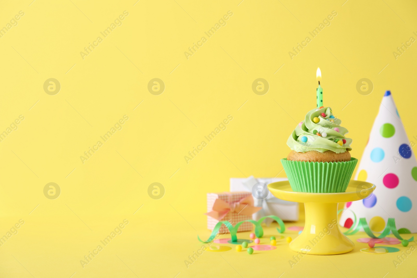 Photo of Delicious birthday cupcake with burning candle, party decor and gift boxes on yellow background, space for text