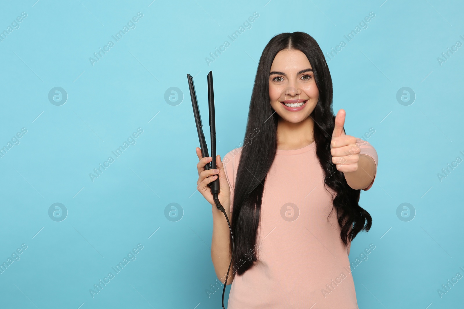 Photo of Beautiful happy woman with hair iron showing thumbs up on light blue background. Space for text