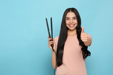 Beautiful happy woman with hair iron showing thumbs up on light blue background. Space for text