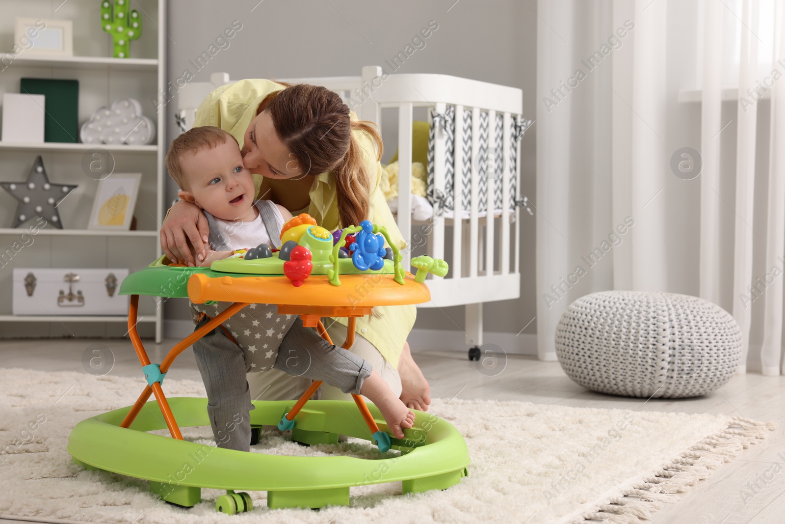 Photo of Mother kissing her little son at home. Cute baby making first steps with toy walker, space for text