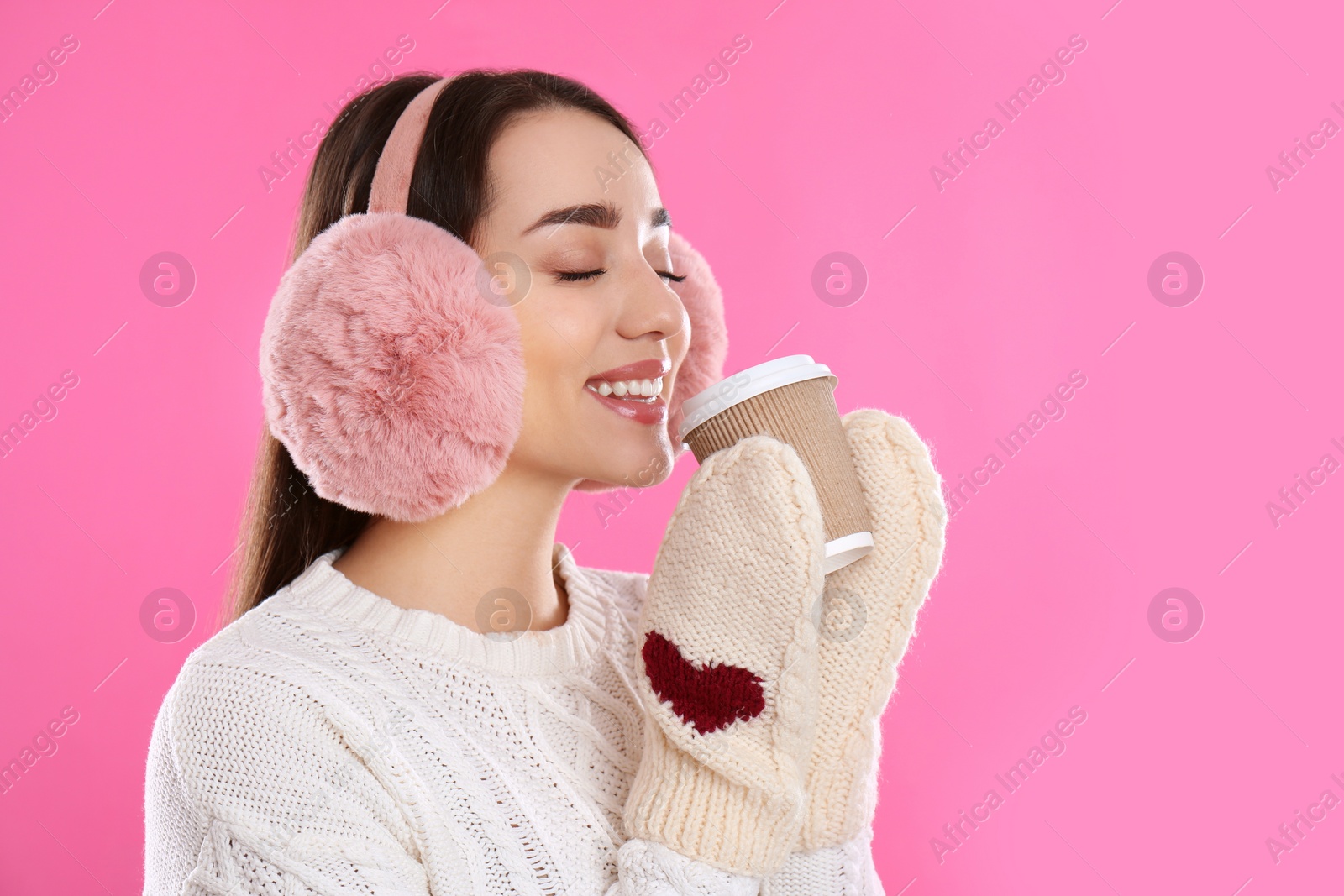 Photo of Beautiful young woman in earmuffs with cup of drink on pink background