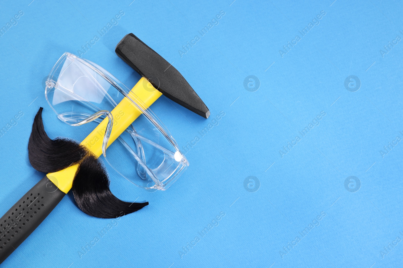 Photo of Man's face made of artificial mustache, safety glasses and hammer on blue background, top view. Space for text