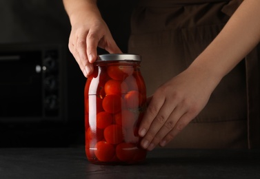 Photo of Woman pickling glass jar of tomatoes at black kitchen table, closeup