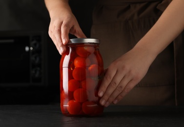 Woman pickling glass jar of tomatoes at black kitchen table, closeup