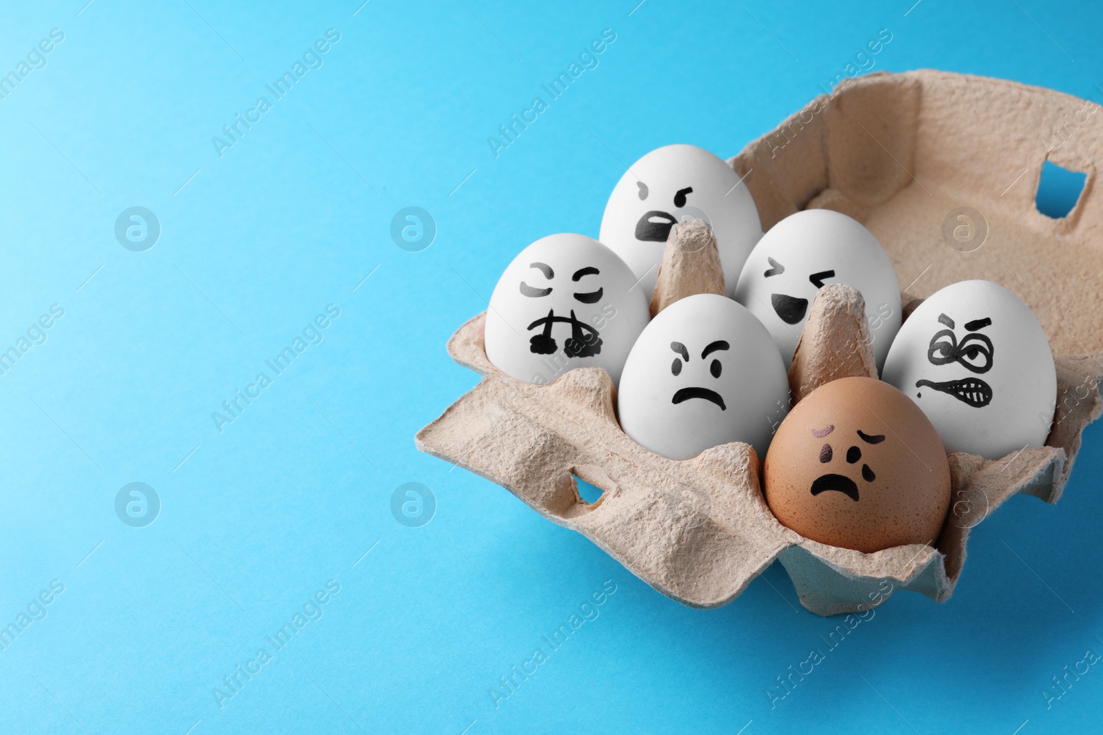 Photo of Brown egg with upset face among aggressively disposed white ones in carton box on turquoise background, space for text. Bullying concept
