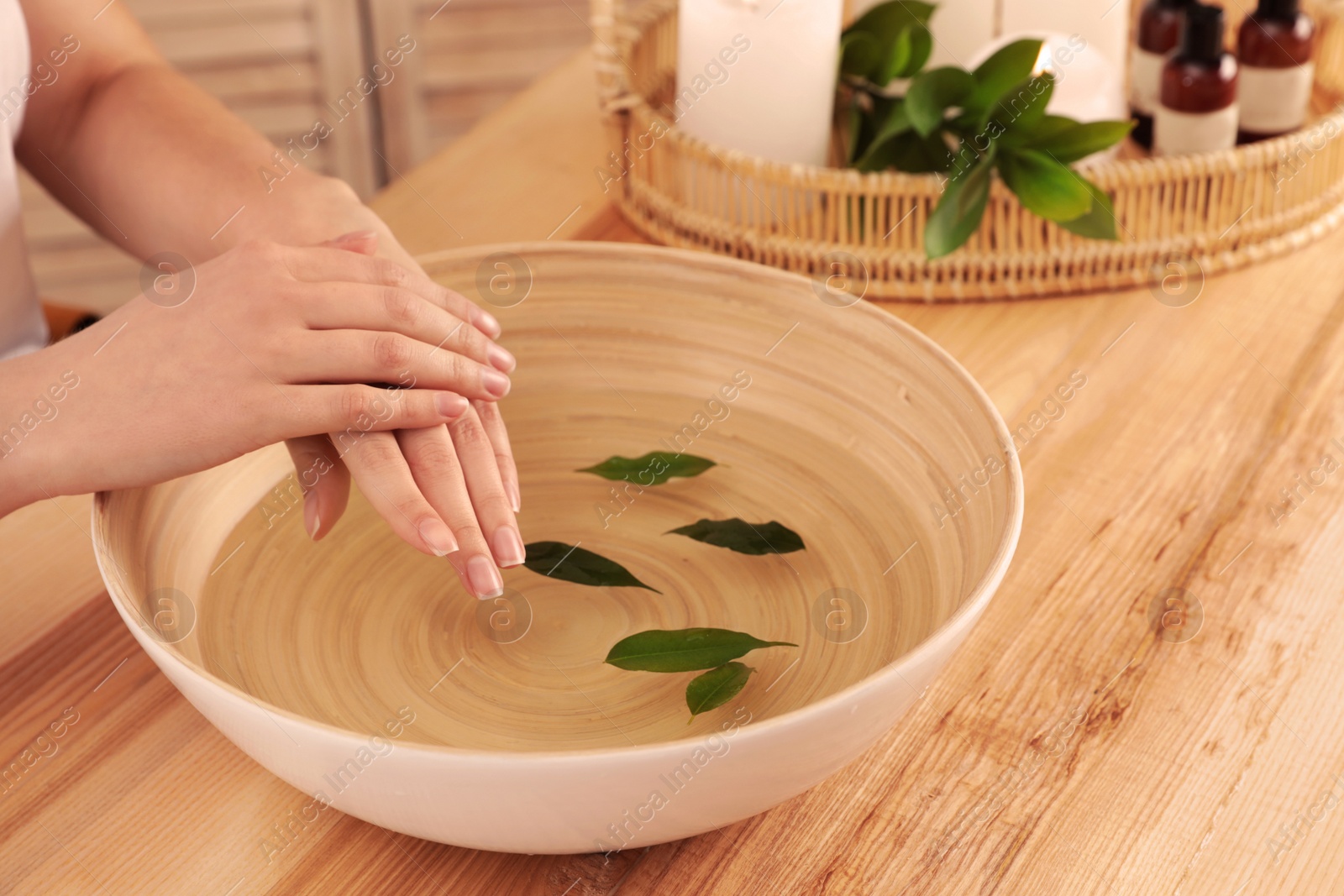 Photo of Woman soaking her hands in bowl of water and leaves on wooden table, closeup with space for text. Spa treatment