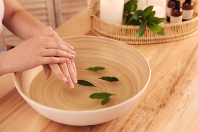 Photo of Woman soaking her hands in bowl of water and leaves on wooden table, closeup with space for text. Spa treatment