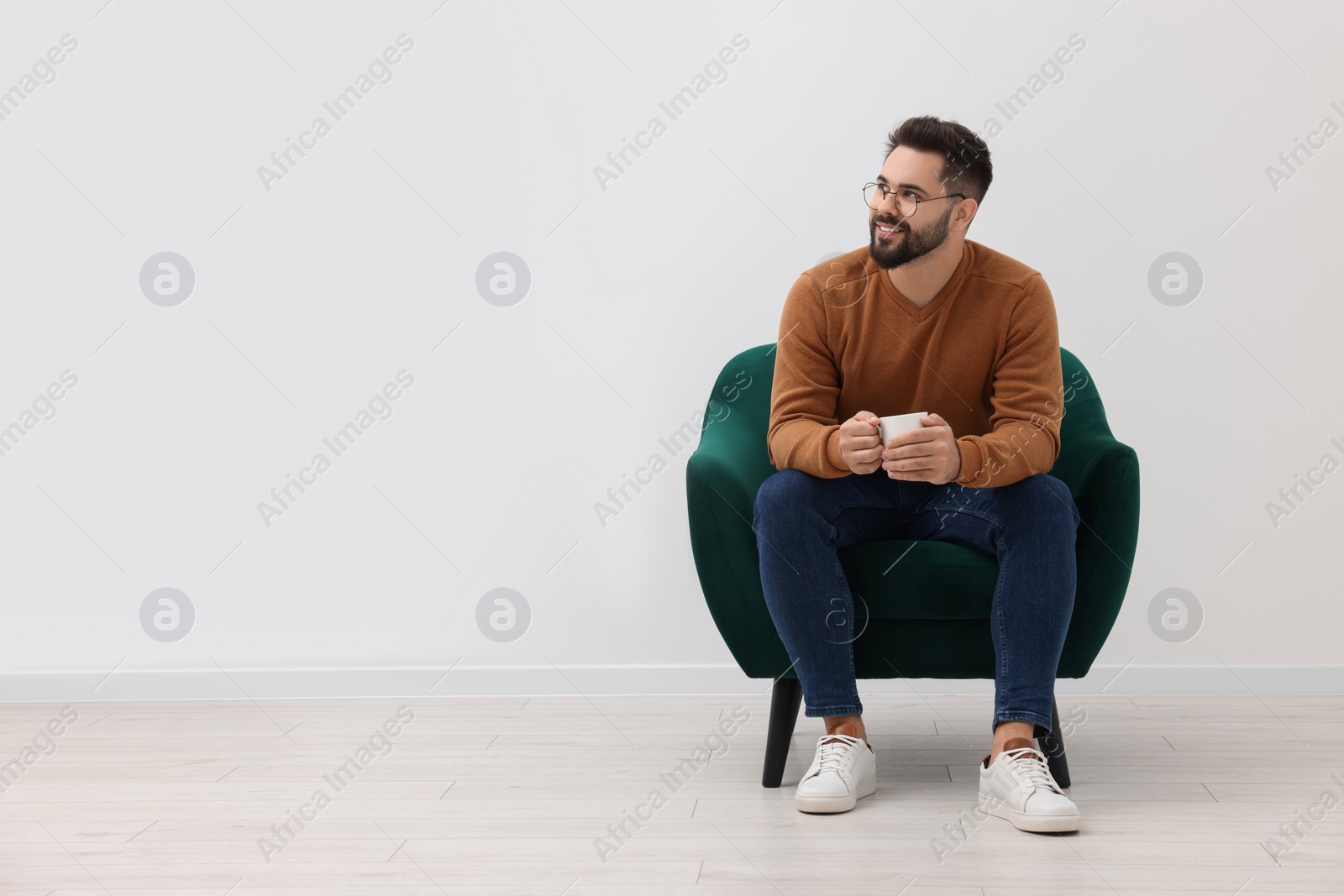 Photo of Handsome man with cup of drink sitting in armchair near white wall indoors, space for text