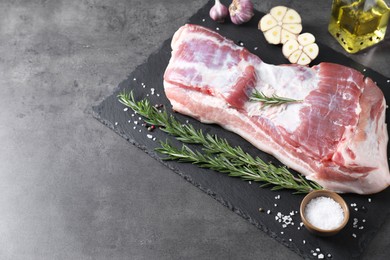 Photo of Piece of raw pork belly, rosemary, garlic and salt on grey textured table, above view. Space for text