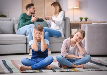 Photo of Little unhappy children sitting on floor while parents arguing at home
