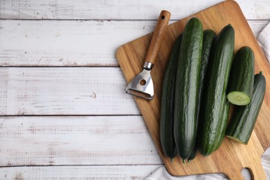 Fresh cucumbers and peeler on white wooden table, top view. Space for text