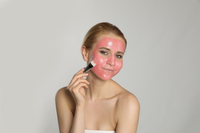 Young woman applying pomegranate face mask on light grey background