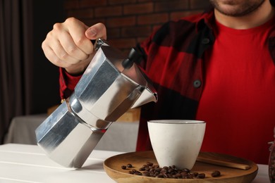 Man pouring aromatic coffee from moka pot into cup at white wooden table indoors, closeup