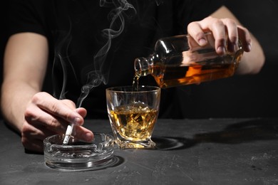 Photo of Alcohol addiction. Man with smoldering cigarette pouring whiskey into glass at dark textured table, closeup