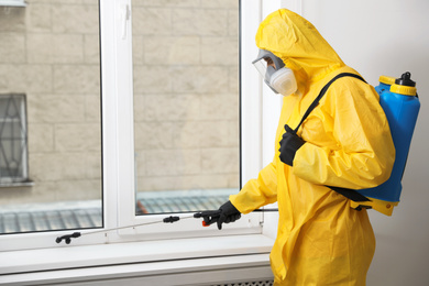 Photo of Pest control worker in protective suit spraying insecticide on window sill indoors. Space for text