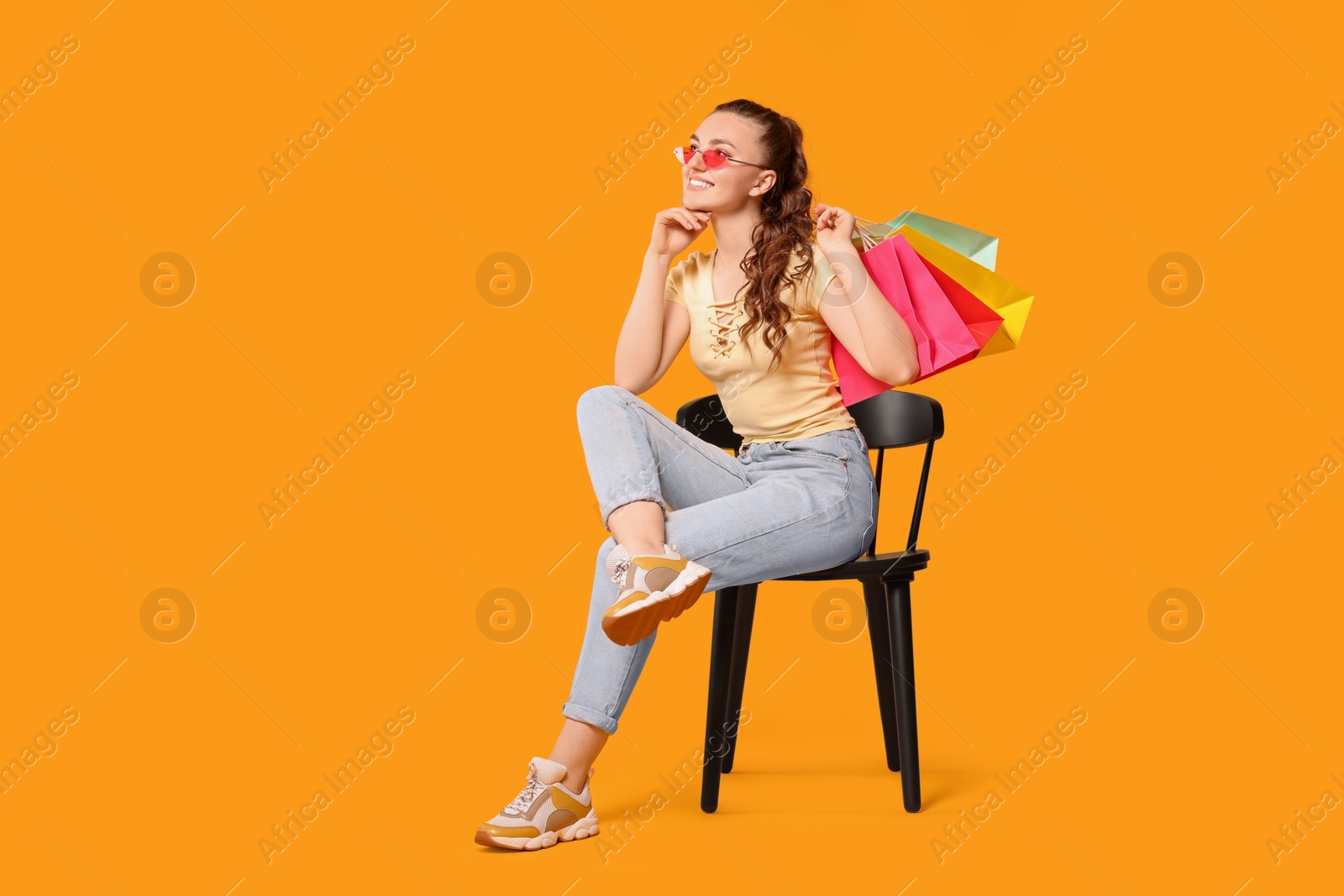 Photo of Happy woman in stylish sunglasses holding colorful shopping bags on chair against orange background