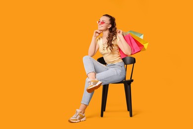 Photo of Happy woman in stylish sunglasses holding colorful shopping bags on chair against orange background