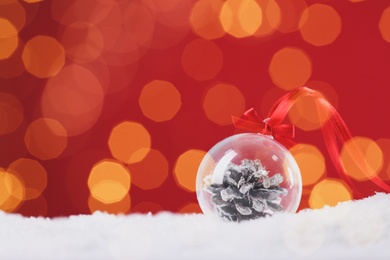 Image of Beautiful transparent Christmas ornament with fir cone on snow against red background, bokeh effect. Space for text. Space for text