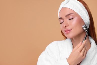 Young woman massaging her face with metal roller on pale orange background, space for text