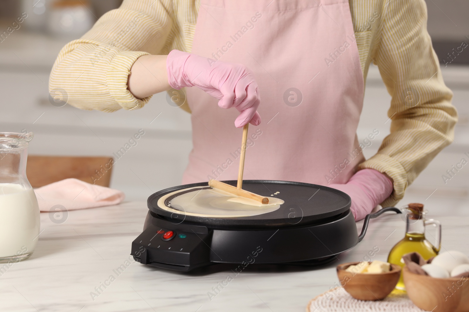 Photo of Woman cooking delicious crepe on electric pancake maker at white marble table in kitchen, closeup