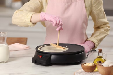 Photo of Woman cooking delicious crepe on electric pancake maker at white marble table in kitchen, closeup