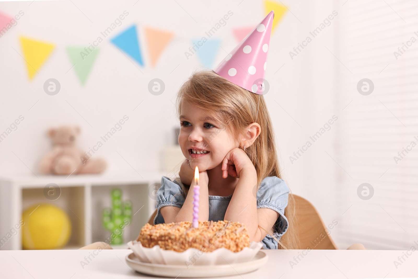 Photo of Cute girl in party hat with birthday cake at table indoors