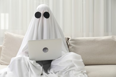 Photo of Creepy ghost. Person covered with white sheet using laptop on sofa at home