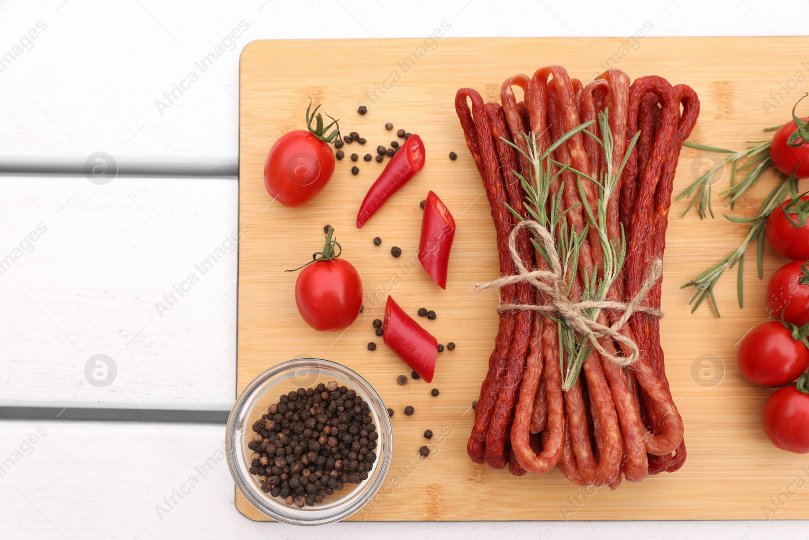 Photo of Bundle of delicious kabanosy with pepper, tomatoes and rosemary on white wooden table, top view. Space for text