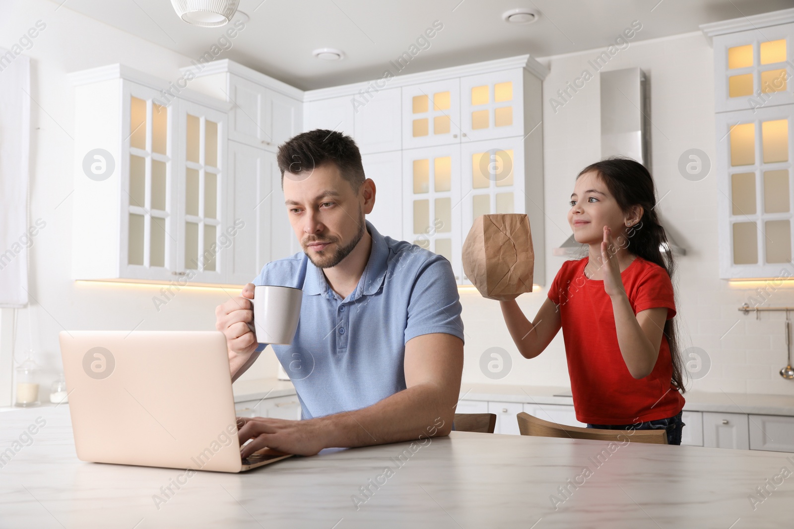 Photo of Cute little girl popping paper bag behind father's back while he working at home