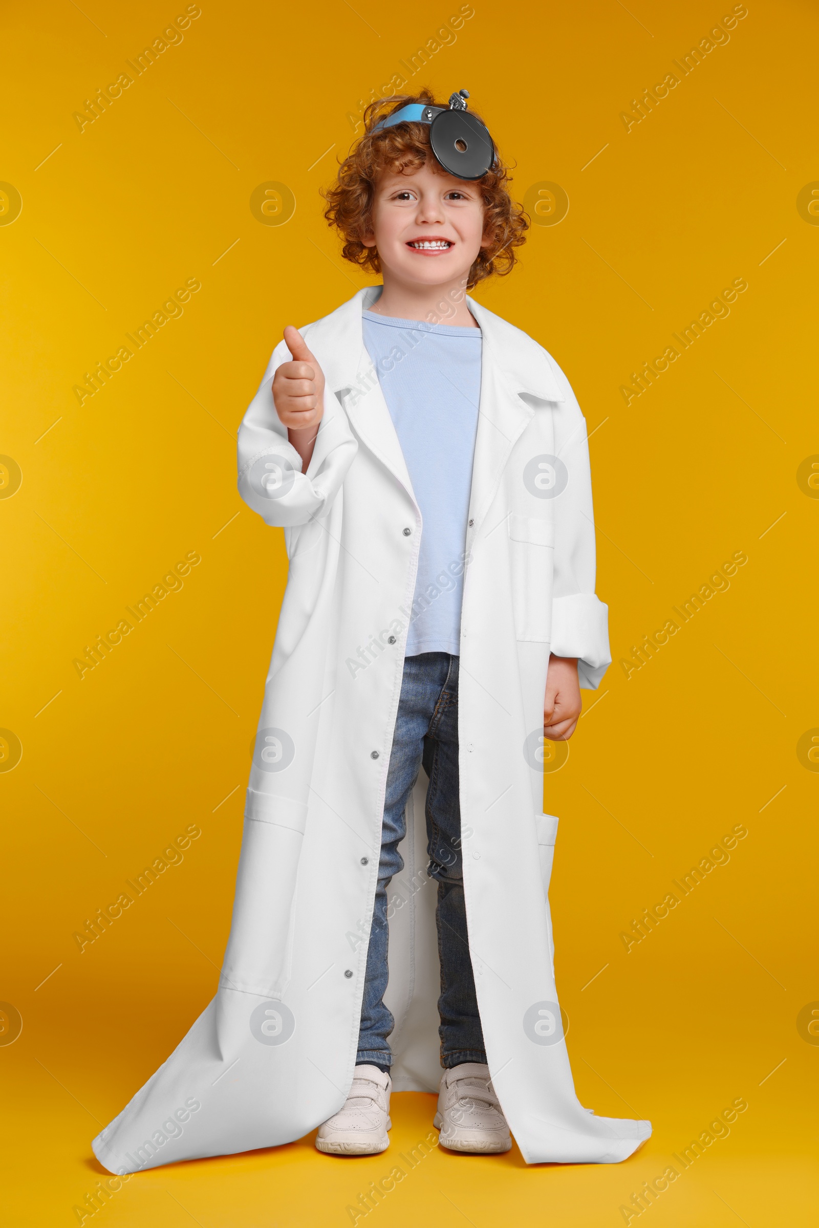 Photo of Little boy in medical uniform with head mirror showing thumb up on yellow background