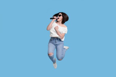 Beautiful young woman with microphone singing and jumping on light blue background