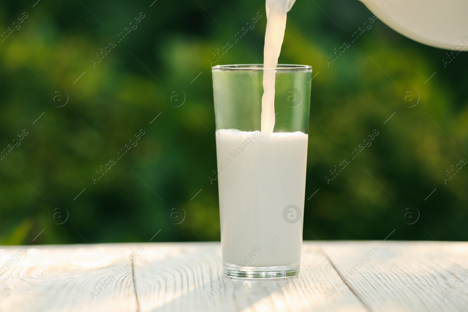 Photo of Pouring tasty fresh milk into glass on white wooden table, closeup. Space for text