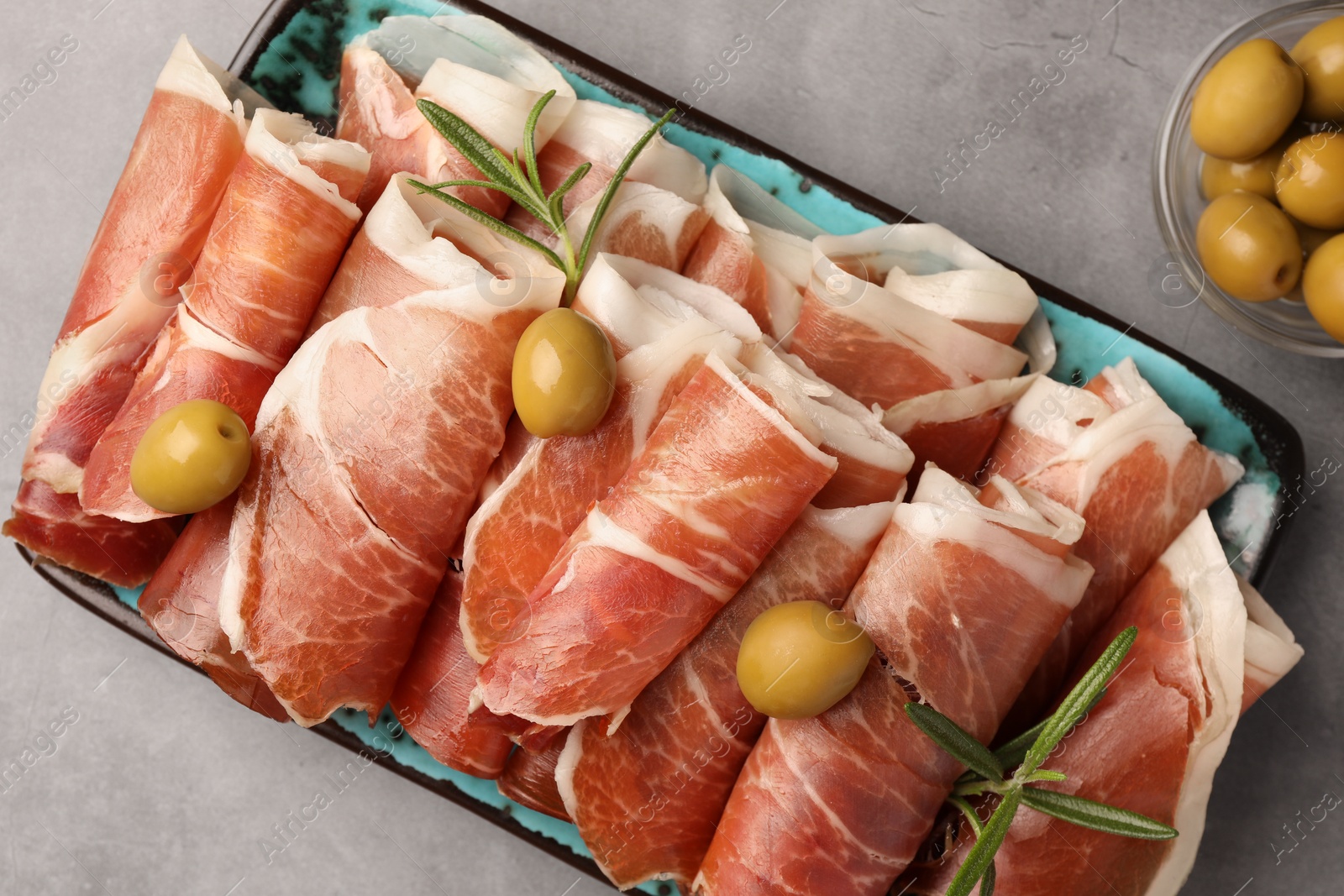 Photo of Rolled slices of delicious jamon with rosemary and olives on grey table, flat lay