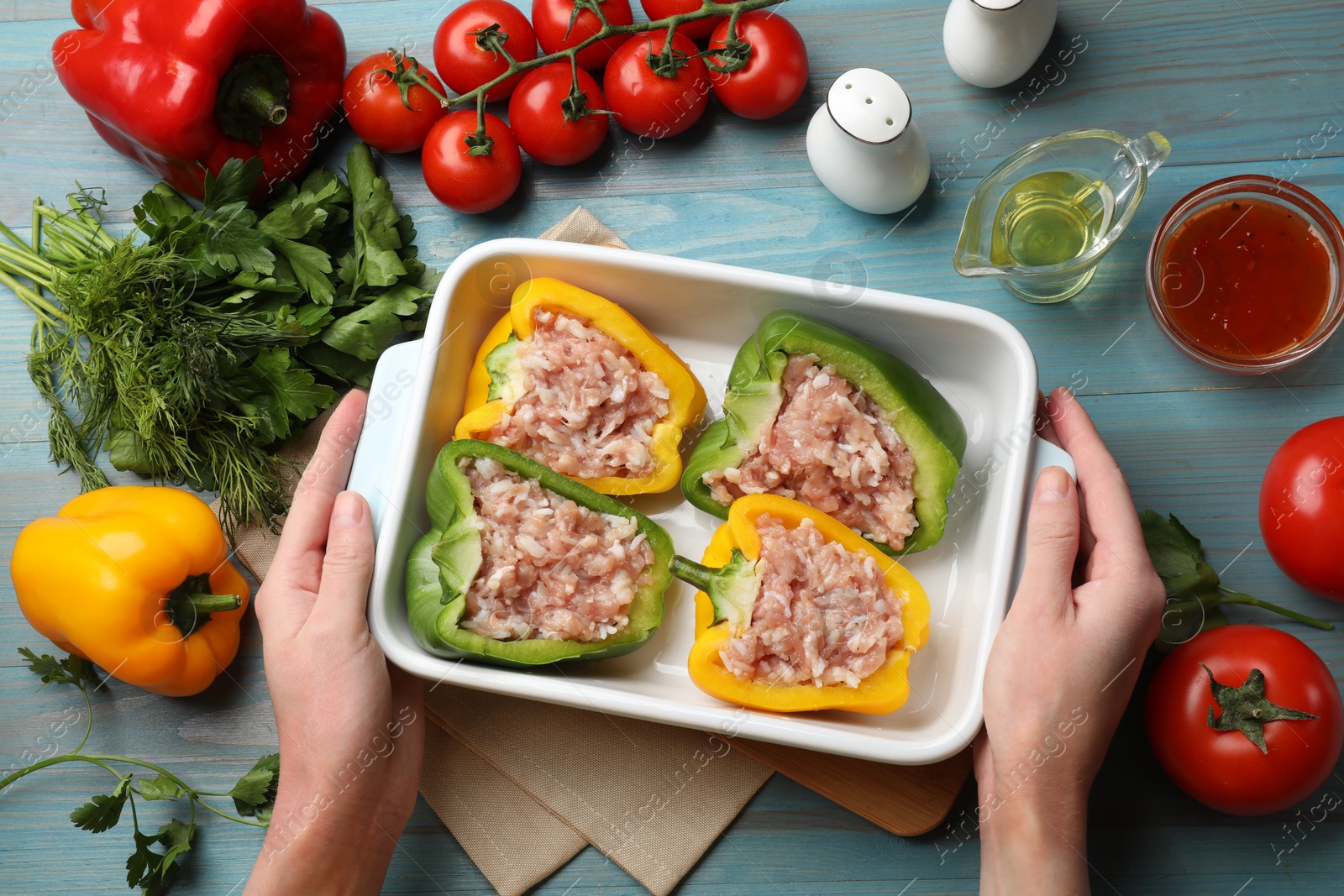 Photo of Woman holding dish with raw stuffed peppers at light blue wooden table, top view