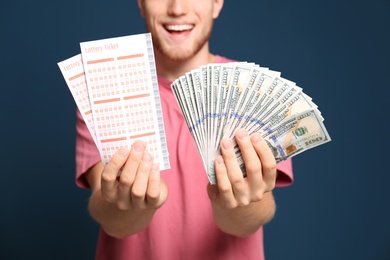 Happy young man holding lottery tickets and money on blue background, closeup