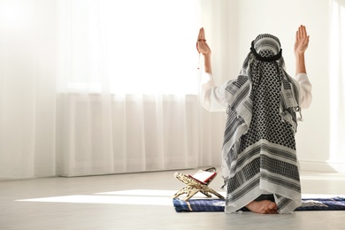 Muslim man in traditional clothes praying on rug indoors. Space for text