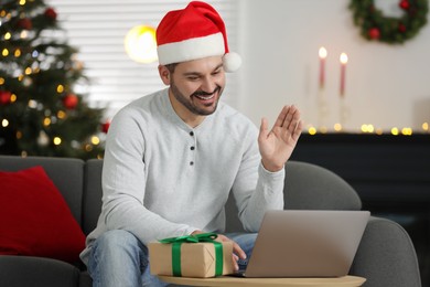 Photo of Celebrating Christmas online with exchanged by mail presents. Smiling man in Santa hat with gift waving hello during video call on laptop at home