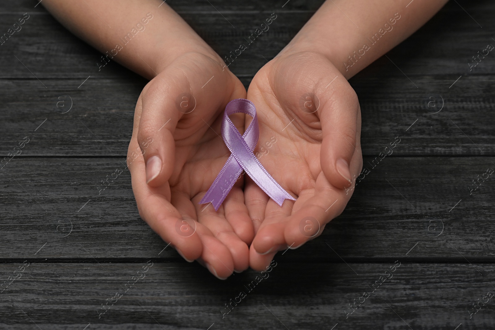 Photo of Woman holding purple awareness ribbon on black wooden background, closeup