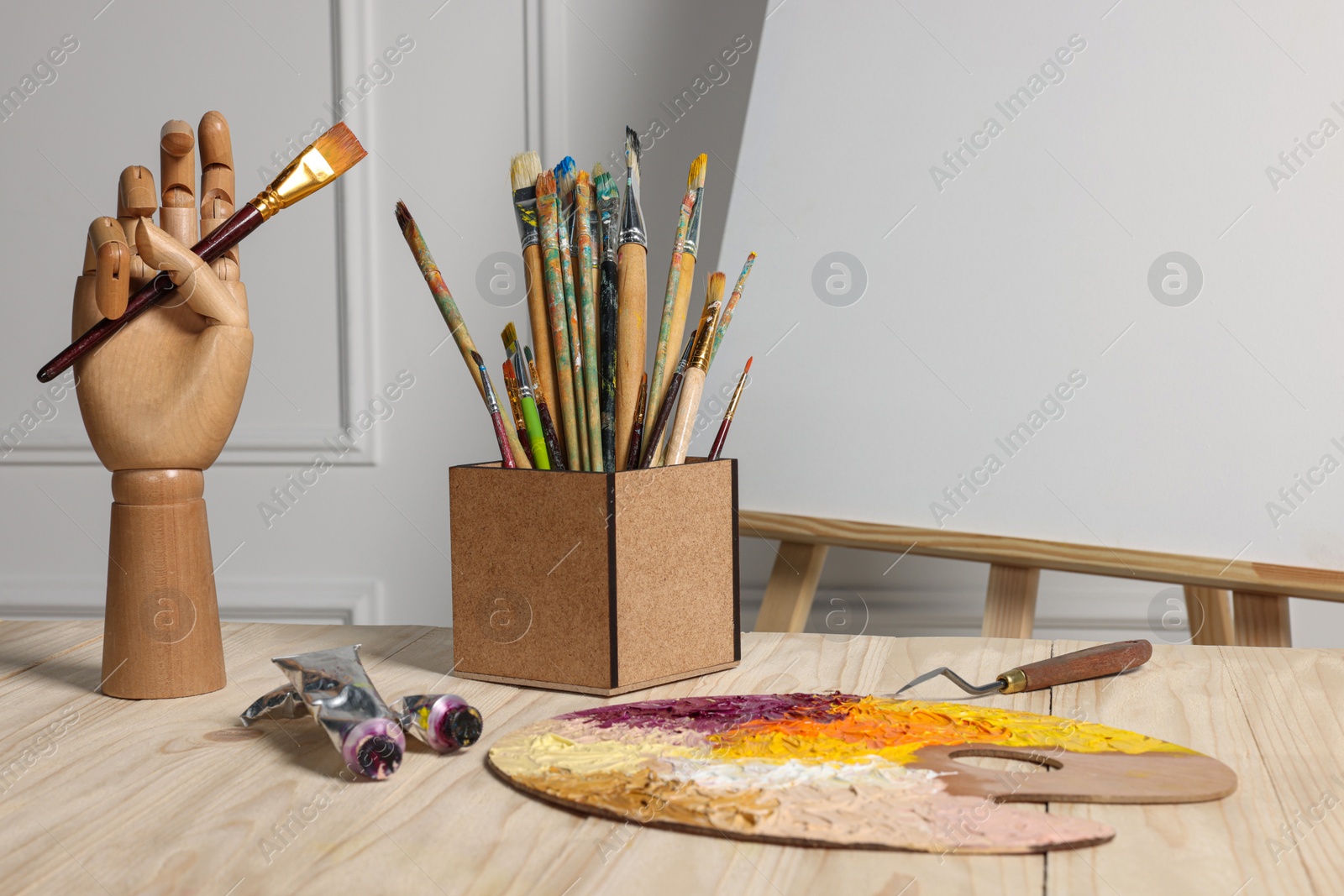 Photo of Easel with blank canvas, hand model and different art supplies on wooden table near white wall